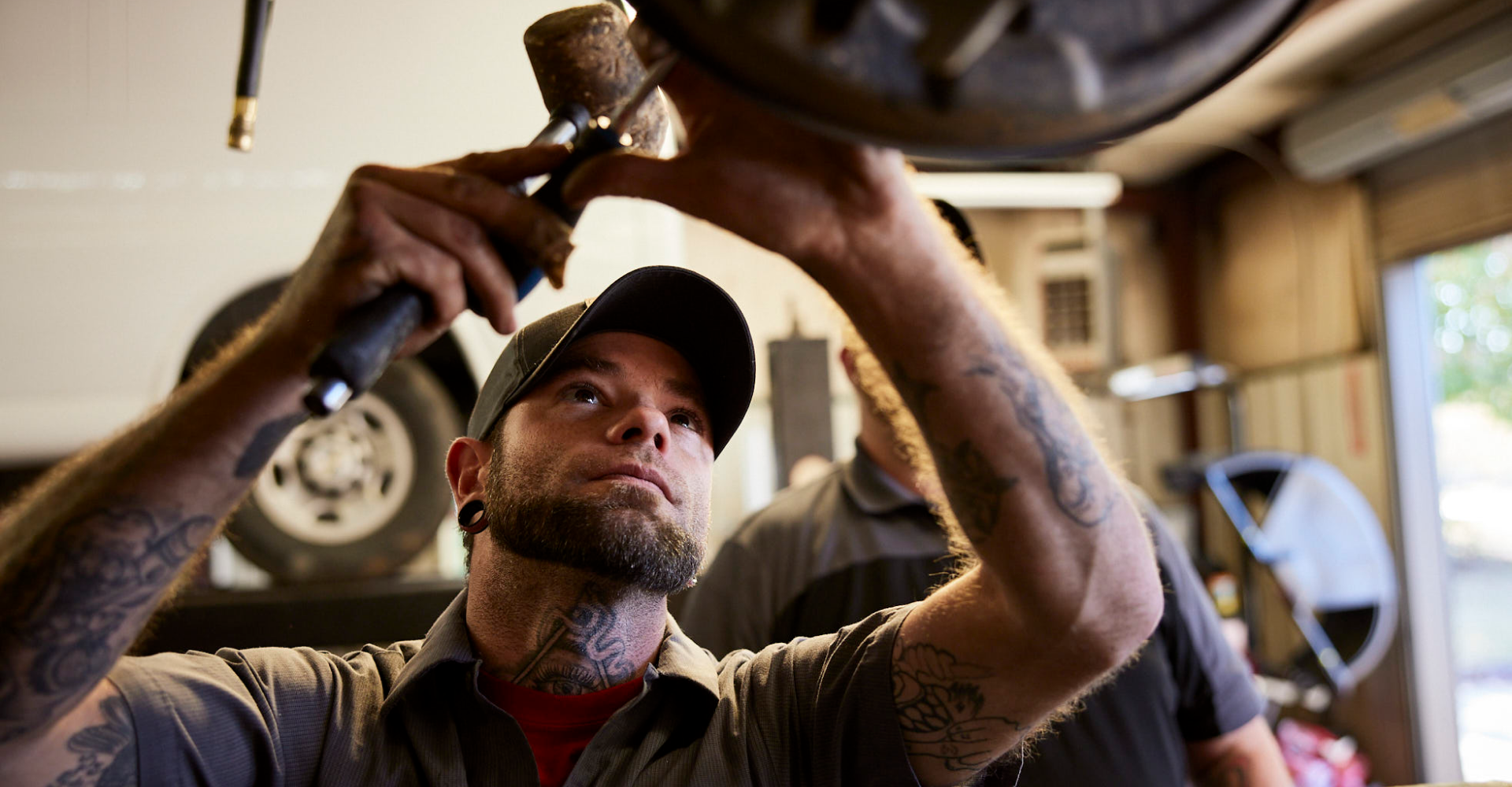 Mechanic wearing a cap, working on a vehicle part under a shop light.