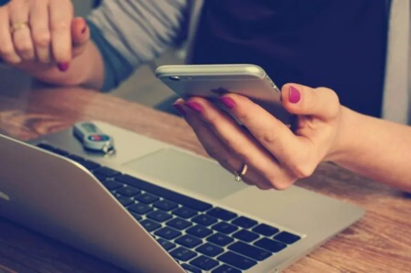 Close‐up of a person’s hand using a smartphone, laptop in the background.